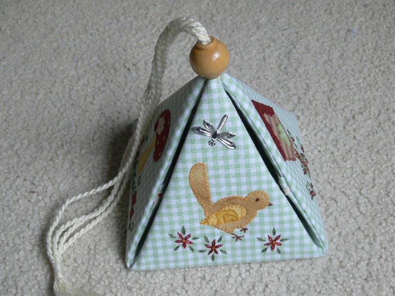 Peut inclure: Une bo&icirc;te en forme de pyramide en tissu vichy vert et blanc avec un cordon blanc et un bouton en bois. La bo&icirc;te est d&eacute;cor&eacute;e de d&eacute;tails brod&eacute;s d'oiseaux, de fleurs et d'une libellule.