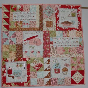 Op de afbeelding: Een patchworkdeken in rood, wit en blauw. De deken heeft verschillende geborduurde ontwerpen met een bakthema, waaronder "Baking Day", "Cook with Love" en afbeeldingen van een deegroller, een taart en cupcakes.