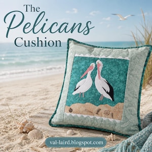 May include: A square decorative cushion featuring a central teal panel with an appliqué of two white pelicans with pink beaks and black wing details. Below the birds is a sandy beige section with small seashell motifs and scalloped borders.