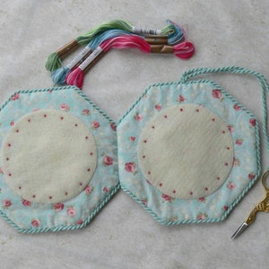 May include: Two hexagonal fabric pin cushions featuring a light blue background with a pink rose pattern, trimmed with twisted mint green cord. Each cushion has a central cream-colored felt circle with small red dots, embroidery floss, and gold scissors.