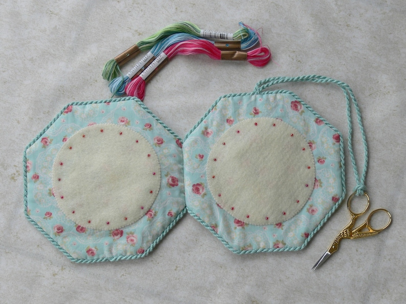 May include: Two hexagonal fabric pin cushions featuring a light blue background with a pink rose pattern, trimmed with twisted mint green cord. Each cushion has a central cream-colored felt circle with small red dots, embroidery floss, and gold scissors.
