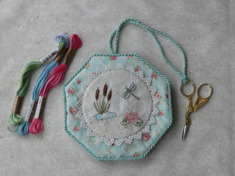 May include: A hexagonal fabric pincushion with a light blue floral pattern, featuring a central circular embroidery of cattails, a dragonfly, and a pink water lily, bordered by white lace. Includes three embroidery floss skeins and gold scissors.