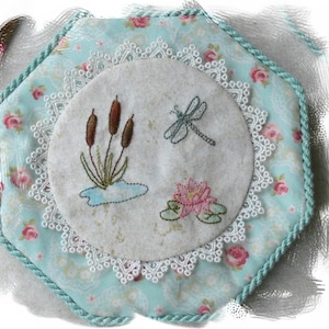 May include: A hexagonal fabric pincushion featuring a light blue floral print with pink roses, bordered by a teal twisted cord. The center displays a circular beige linen panel with white lace trim, embroidered with brown cattails, a dragonfly, and a pink lotus.