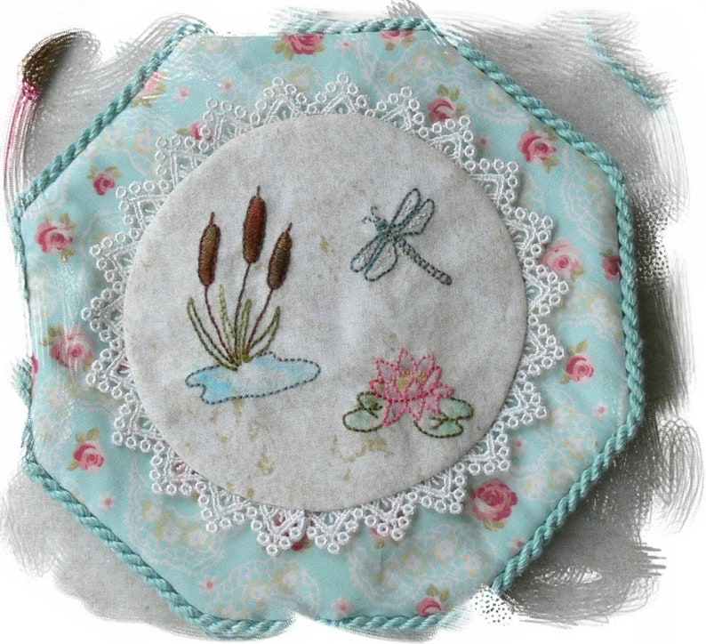 May include: A hexagonal fabric pincushion featuring a light blue floral print with pink roses, bordered by a teal twisted cord. The center displays a circular beige linen panel with white lace trim, embroidered with brown cattails, a dragonfly, and a pink lotus.