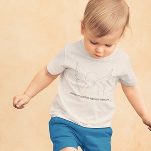 May include: A young child wearing a white t-shirt with a line drawing of an angel with wings and the text "making my guardian angel work overtime." The child is wearing blue shorts and brown shoes and is kicking a black and white football.