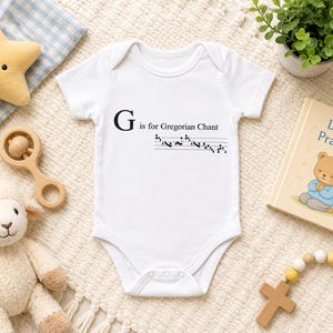 May include: White baby onesie with the text "G is for Gregorian Chant" and musical notes. A plush star, wooden rattle, stuffed lamb, small potted plant, and a "Little Prayers" book are also visible.