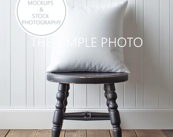 May include: A white pillow sits on a black wooden stool with turned legs. The stool is on a wood floor in front of a white paneled wall.