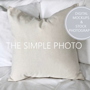 May include: A white pillow with a plain, off-white fabric cover. The pillow is lying on a white bed with other pillows behind it. The text "THE SIMPLE PHOTO" is printed on the bedspread. A gray circle with the text "DIGITAL MOCKUPS & STOCK PHOTOGRAPHY" is in the upper right corner of the image.