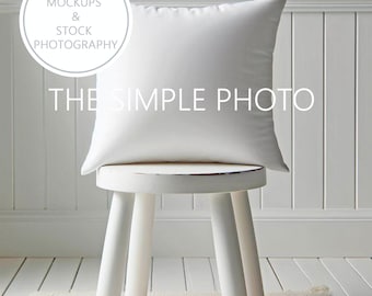 May include: A square white pillow rests on a small, white, three-legged stool. The stool is placed on a light-colored rug. The background is a white wall with vertical paneling. Text on the image reads "DIGITAL MOCKUPS & STOCK PHOTOGRAPHY" and "THE SIMPLE PHOTO."
