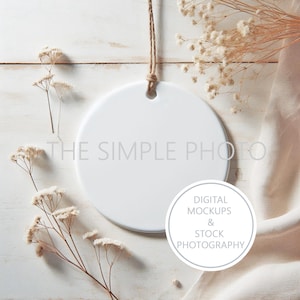 May include: A white ceramic ornament with a string hanging from the top. The ornament is on a white wooden surface with dried flowers and a white fabric.