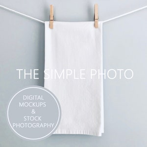 May include: A white kitchen towel hanging on a clothesline with wooden clothespins. The towel is blank and ready for customization. The text "THE SIMPLE PHOTO" is printed below the towel. The text "DIGITAL MOCKUPS & STOCK PHOTOGRAPHY" is printed in a circle below the text.