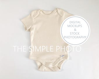 May include: A plain, off-white baby onesie with short sleeves. The onesie is laid flat on a white background. The text "THE SIMPLE PHOTO" is visible at the bottom of the image. A circle with the text "DIGITAL MOCKUPS & STOCK PHOTOGRAPHY" is visible in the top right corner of the image.