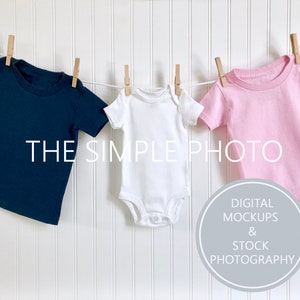 Op de afbeelding: Drie lege baby rompertjes hangen aan een waslijn tegen een witte houten paneel achtergrond. De rompertjes zijn donkerblauw, wit en roze. De tekst "THE SIMPLE PHOTO" is in wit op de achtergrond gedrukt. Onder de rompertjes bevindt zich een grijze cirkel met de tekst "DIGITAL MOCKUPS & STOCK PHOTOGRAPHY".