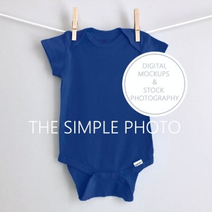 May include: A royal blue baby onesie hanging on a clothesline with clothespins. The onesie is blank and ready for customization. The text "DIGITAL MOCKUPS & STOCK PHOTOGRAPHY" is in a circle on the right side of the image. The text "THE SIMPLE PHOTO" is below the onesie.