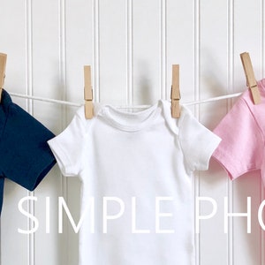 Op de afbeelding: Drie blanco t-shirts hangen aan een waslijn met houten wasknijpers. De shirts zijn donkerblauw, wit en roze.