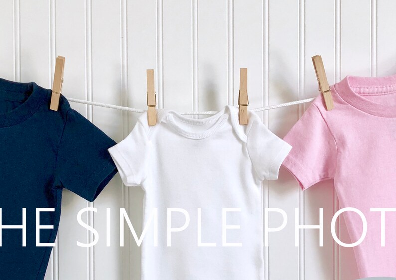 Op de afbeelding: Drie blanco t-shirts hangen aan een waslijn met houten wasknijpers. De shirts zijn donkerblauw, wit en roze.