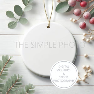 May include: White ceramic ornament with a gold string hanging from the top. The ornament is blank and ready to be decorated. The ornament is surrounded by white wood and green leaves.