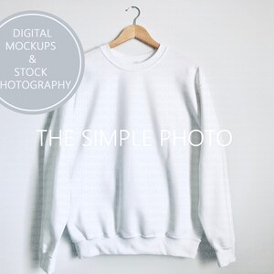 May include: A plain white sweatshirt hangs on a wooden hanger against a white background. The text "DIGITAL MOCKUPS & STOCK PHOTOGRAPHY" is in a gray circle in the upper left. The words "THE SIMPLE PHOTO" are in white across the center of the sweatshirt.
