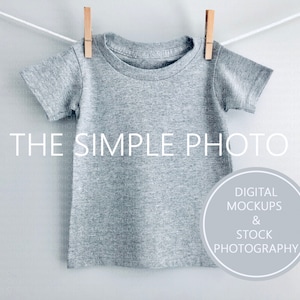 Może przedstawiać: Szary T-shirt z krótkim rękawem wiszący na sznurku do suszenia z drewnianymi klamrami. Koszulka jest pusta i gotowa do personalizacji. Tekst "THE SIMPLE PHOTO" jest widoczny na dole obrazu. Koło z tekstem "DIGITAL MOCKUPS & STOCK PHOTOGRAPHY" jest widoczne w prawym dolnym rogu.