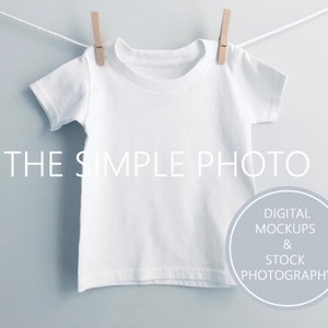 Może przedstawiać: Biała koszulka wisząca na sznurze do suszenia z drewnianymi klamrami. Koszulka jest pusta i gotowa do personalizacji. Tekst "THE SIMPLE PHOTO" jest wydrukowany pod koszulką. Tekst "DIGITAL MOCKUPS & STOCK PHOTOGRAPHY" jest wydrukowany w kółku pod tekstem.