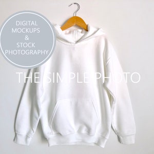 Może przedstawiać: Biała bluza z kapturem z przednią kieszenią wisi na drewnianym wieszaku na białej ścianie. Bluza jest prosta, z tekstem "THE SIMPLE PHOTO" i "DIGITAL MOCKUPS & STOCK PHOTOGRAPHY" w tle.