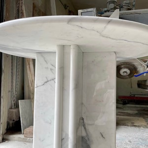 May include: A white marble table with a round top and a rectangular base. The table has gray veining throughout the marble. The base has three vertical, rounded columns. The table is in a workshop setting.