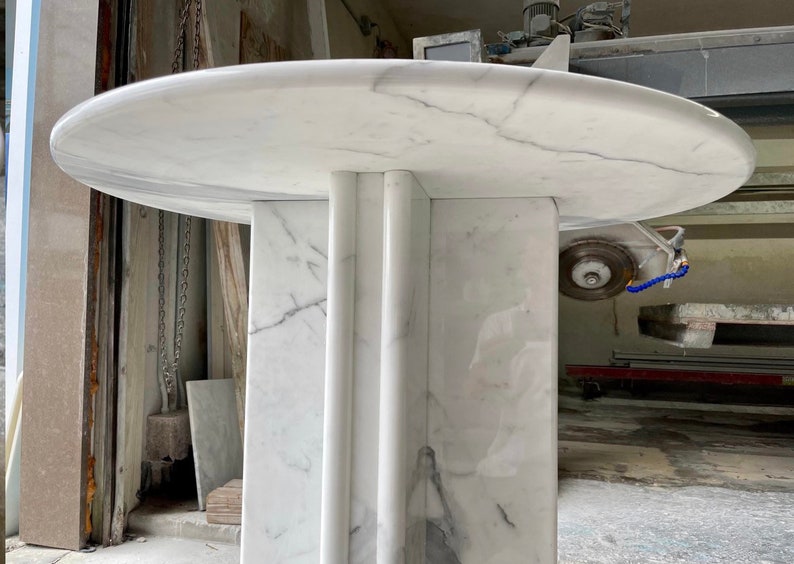 May include: A white marble table with a round top and a rectangular base. The table has gray veining throughout the marble. The base has three vertical, rounded columns. The table is in a workshop setting.