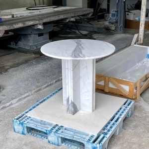 May include: A round marble table with a white and gray veined top. The pedestal base is a geometric design with vertical panels. The table is displayed on a blue plastic pallet in a workshop setting.