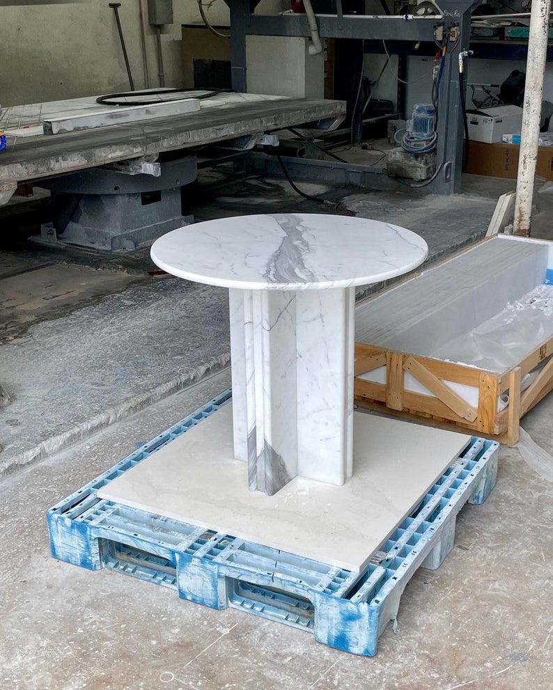 May include: A round marble table with a white and gray veined top. The pedestal base is a geometric design with vertical panels. The table is displayed on a blue plastic pallet in a workshop setting.