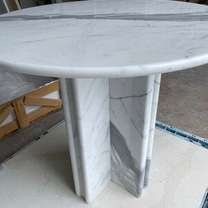 May include: A round marble table with a white surface and gray veining. The table has a pedestal base with a cross-shaped design. The table is approximately 40 inches in diameter.