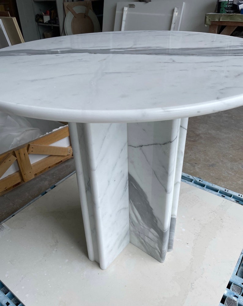 May include: A round marble table with a white surface and gray veining. The table has a pedestal base with a cross-shaped design. The table is approximately 40 inches in diameter.