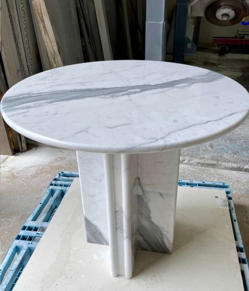 May include: A round marble table with a white and gray veined top. The table has a pedestal base with a cross-shaped design. The table is approximately 30 inches in diameter.