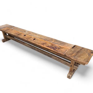 May include: A long, narrow wooden bench featuring a weathered, natural brown finish with visible wood grain, knots, and imperfections. The bench is supported by two sturdy, rectangular trestle-style legs connected by a horizontal support beam.