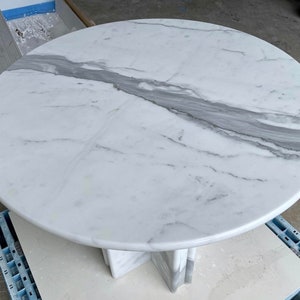 May include: A round white marble table with gray veining. The table has a pedestal base. The marble top is smooth and polished. The table is suitable for dining or as a decorative accent piece.