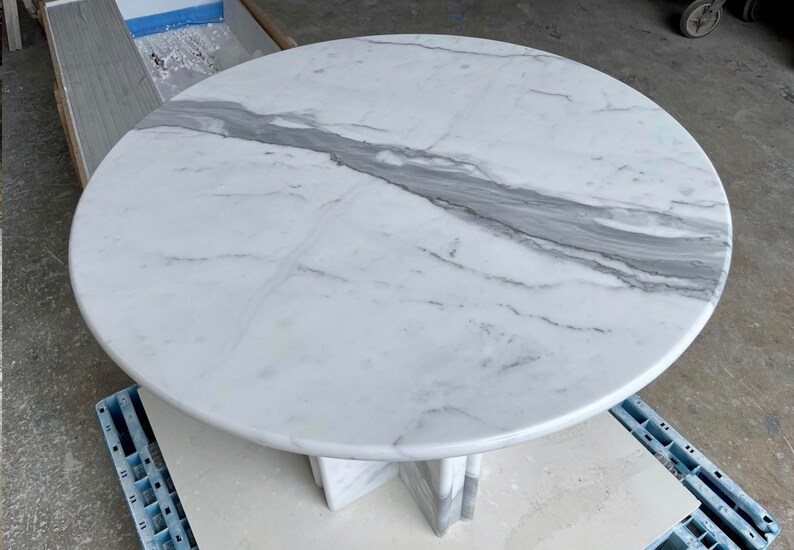 May include: A round white marble table with gray veining. The table has a pedestal base. The marble top is smooth and polished. The table is suitable for dining or as a decorative accent piece.