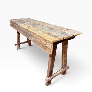 May include: A long, narrow bench constructed from weathered, reclaimed wood with a natural brown and grey finish. The bench features a thick, rectangular top surface and sturdy, angled legs connected by horizontal support beams at the base.