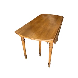 May include: A light brown, oval-shaped drop-leaf table with four turned legs. The table has a wooden surface and brass accents on the feet. The table is in a classic style.