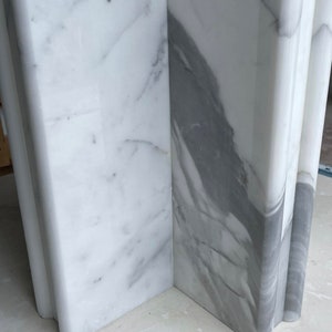 May include: Close-up of a section of a white marble structure with gray veining. The marble has a polished finish and is cut into a rectangular shape with rounded edges. The veining creates a natural pattern.
