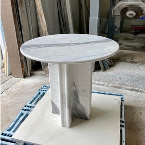 May include: A round marble table with a white and gray veined top. The pedestal base is a matching marble with a geometric design. The table is sitting on a blue pallet.