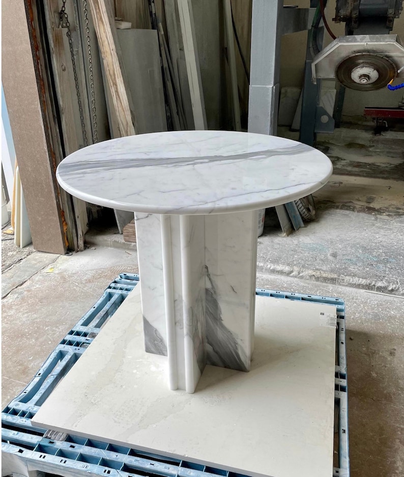 May include: A round marble table with a white and gray veined top. The pedestal base is a matching marble with a geometric design. The table is sitting on a blue pallet.