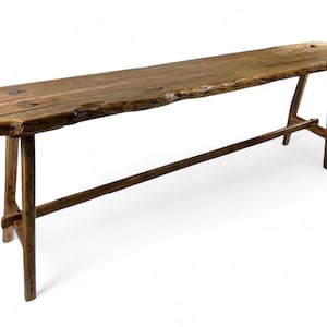 May include: A long, narrow wooden bench featuring a natural live edge top with a dark brown finish. The bench is supported by four angled legs connected by a horizontal stretcher bar, showcasing a rustic, weathered texture and visible wood grain.