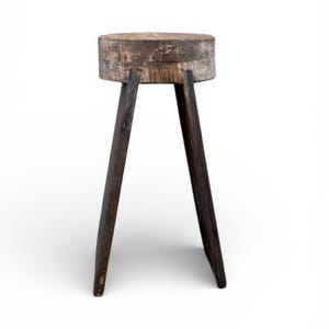 May include: A rustic wooden stool with a circular top and three dark brown legs. The top appears to be a cross-section of a tree trunk, showcasing the natural wood grain. The legs are angled for stability.