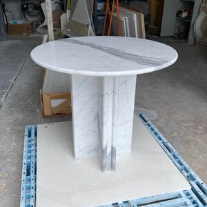 May include: A round marble table with a white and gray veined top. The table has a pedestal base with vertical accents. The table is approximately 36 inches in diameter and stands on a blue pallet.