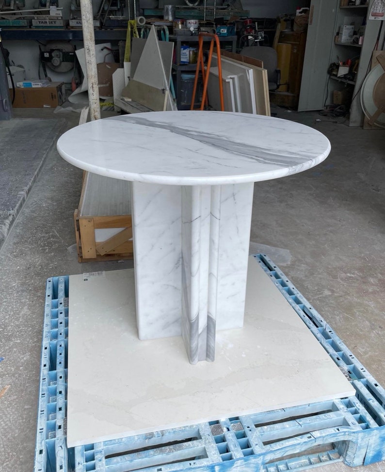 May include: A round marble table with a white and gray veined top. The table has a pedestal base with vertical accents. The table is approximately 36 inches in diameter and stands on a blue pallet.