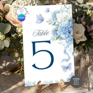 May include: A white table number card with a blue floral design and butterflies. The card features the number 5 in a dark blue font. The text "Table" is above the number. The card is displayed on a gold stand. Available in 10.16 x 15.24 cm, 12.7 x 17.78 cm, and 20.32 x 25.4 cm sizes.
