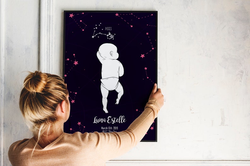 May include: A black framed poster with a dark blue background and pink stars. The poster features a white outline of a baby standing with the constellation Pisces above the baby. The text on the poster reads "Luna Estelle, March 8th, 2019, 8:11 PM, Pisces".