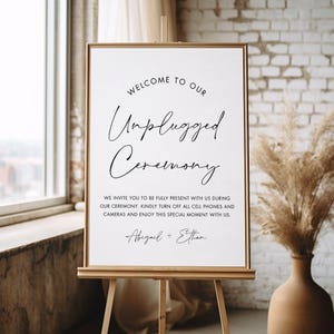 Puede incluir: Un letrero enmarcado sobre un caballete de madera con el texto "Welcome to our Unplugged Ceremony". El letrero también incluye una solicitud para apagar teléfonos y cámaras. El letrero está en una habitación con una ventana y un jarrón.