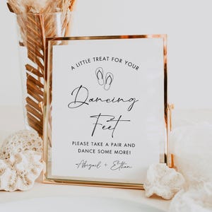 May include: A gold framed sign with the text "A LITTLE TREAT FOR YOUR Dancing Feet" and "PLEASE TAKE A PAIR AND DANCE SOME MORE!" in black script. The sign includes the names "Abigail + Ethan" at the bottom. The sign is decorated with a pair of flip-flops.
