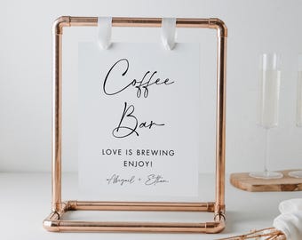 May include: A copper-colored metal sign holder with a white card that reads "Coffee Bar" in elegant script. The sign also includes the text "LOVE IS BREWING ENJOY!" and the names "Abigail + Ethan". White ribbons hold the card in place.