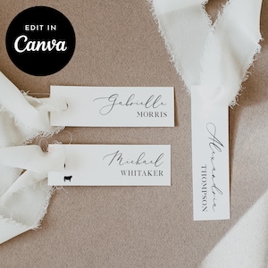 May include: White place cards with elegant calligraphy names, including "Gabrielle Morris" and "Michael Whitaker." A black circle with "Edit in Canva" text is in the upper left. Soft white ribbon accents the cards, suggesting a wedding or event theme.